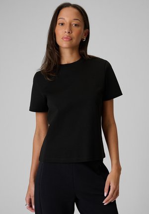 Kobieta o średnim odcieniu skóry i długich ciemnych włosach, ubrana w gładką czarną koszulkę z krótkim rękawem i czarne spodnie na szarym tle.