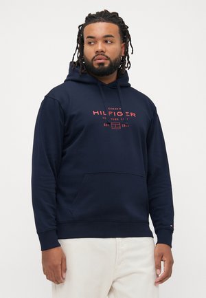 Mann mit Dreadlocks, der einen marineblauen Tommy Hilfiger-Hoodie mit Kängurutasche und weißen Hosen trägt, und nach links schaut.