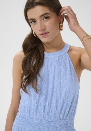 Hosszú barna hajú nő, akinek világoskék csipkés ujjatlan felsője, arany karika fülbevalói és arany karkötői vannak, és a haját igazgatja.