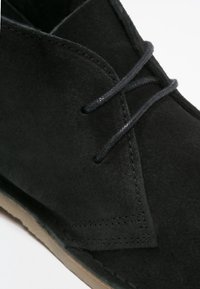 Bottines noires en suède avec une texture lisse, dotées de deux œillets, de lacets noirs et d'une semelle en caoutchouc marron clair.