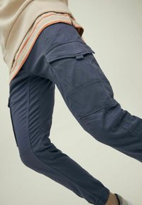 Osoba v námořnických cargo kalhotách a béžových vrstvených tričkách, zvedající jedna noha, ukazující detaily kapes a elastické manžety na kotnících.