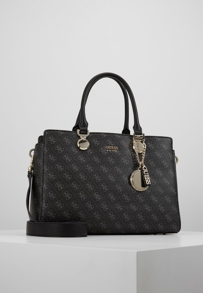 Sac à main noir Guess avec doubles poignées, motif monogramme, logo doré et charms en chaîne, posé sur un socle blanc sur un fond clair uni.