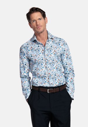 Man draagt een blauw-wit shirt met lange mouwen met bloemenprint, ingestopt in zwarte broek met een bruine riem, handen in de zakken.