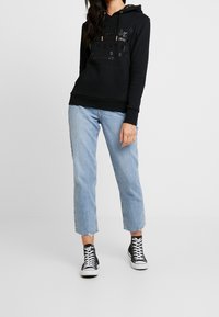 Felpa nera con testo in rilievo, tasca anteriore e lacci. Indossata con jeans dritti blu chiaro e sneakers nere in tela.