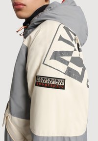 Huvjacka i krämfärg och grått, med en stor grafisk tryck och en svart patch-logotyp. Tillverkad av ett mjukt, möjligtvis vattentätt material.