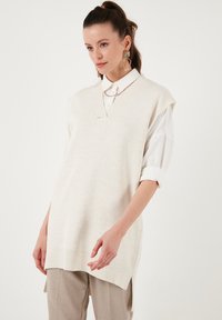Lys beige strikket vest med V-hals, tekstureret stof og slidser i siden, båret over en hvid skjorte med krave. Ingen synlige mønstre eller metaldele.