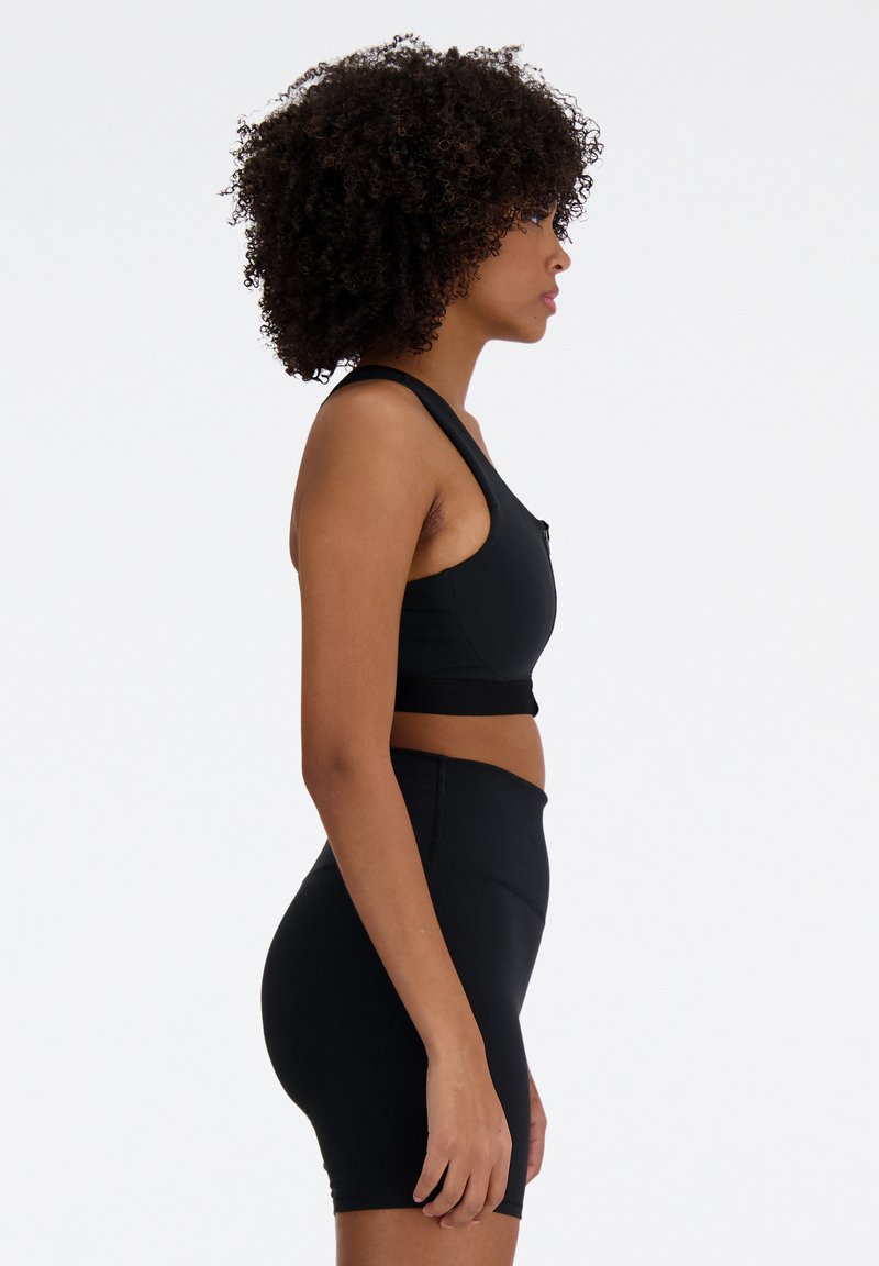 Femme aux cheveux bouclés portant un soutien-gorge de sport noir et un short de vélo taille haute, se tenant de profil sur fond blanc.