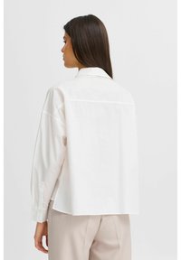 Camicia bianca in cotone over size con maniche lunghe, una cucitura sul dietro e orlo arrotondato, abbinata a pantaloni beige su misura.