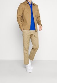 Brun, knappad jacka över en blå polo-shirt, ihop med beige byxor och vita sneakers. Slät tyg och avslappnad passform.