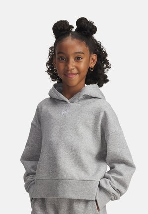 Fille souriante avec des cheveux bouclés coiffés en deux chignons, portant des boucles d'oreilles en or et un hoodie gris clair Under Armour avec les mains dans les poches.
