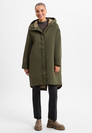 Lang olijfgroene winterjas met capuchon, voorzien van een rits aan de voorkant en drukknoopsluiting, gecombineerd met zwarte broek en beige stevige laarzen.