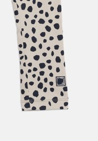 Μανίκι από μπεζ ριγέ ύφασμα με ακανόνιστες μαύρες κηλίδες και ένα μικρό ορθογώνιο κεντημένο λογότυπο σε μαύρο και λευκό κοντά στην μανσέτα.