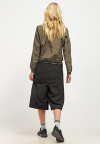 Olijf groene bomberjack met elastische manchetten, gecombineerd met zwarte wijde shorts, casual zwarte sneakers en witte gestreepte sokken. Haar is golvend.