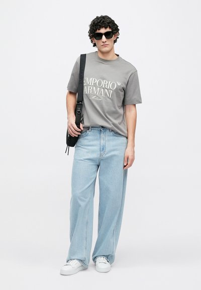 T-shirt cinzenta com o logótipo "Emporio Armani", jeans azul claro de pernas largas, ténis brancos e óculos de sol pretos. Bolsa preta ao ombro visível.