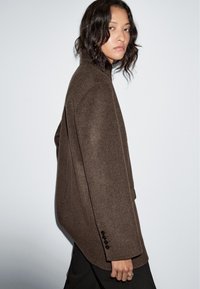 Brown wool blend jacket with a high collar, oversized fit, and four black buttons on the sleeve. Smooth texture, minimalistic design.