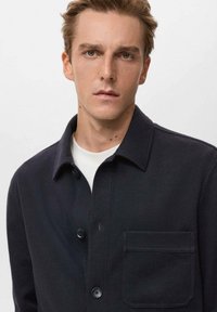 Camicia button-up blu navy in un materiale morbido e testurizzato, con colletto, tasca frontale e design semplice. T-shirt bianca a girocollo visibile.