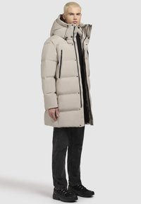 Beige pufferjakke med en høj, justerbar hætte, frontlynlås og sorte lynlåslommer. Bæres over en sort top og parret med mørke jeans.