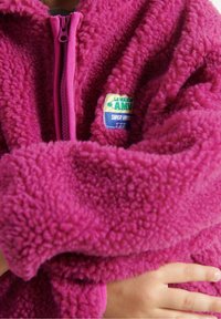 Barn iført en tekstureret lys pink fleecejakke med lynlås foran og et lille farverigt mærke på brystet, med armene korslagt over kroppen.