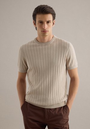 Jonge man met donker haar die een beige geribde korte mouw trui en bruine broek draagt, staand met handen in de zakken tegen een effen achtergrond.