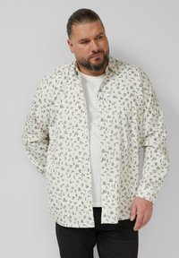 Witte overhemd met zwarte patronen van verschillende kleine iconen, met een klassieke kraag en lange mouwen, gedragen over een wit T-shirt.