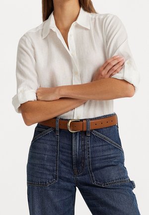 Femme portant une chemise blanche boutonnée aux manches retroussées, un jean bleu taille haute et une ceinture en cuir marron avec boucle dorée, bras croisés.