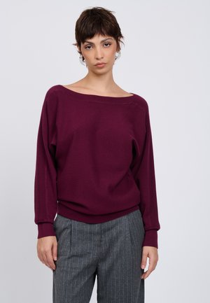 Junge Frau mit kurzen dunklen Haaren, die einen lockeren bordeauxfarbenen Pullover und graue Nadelstreifenhosen trägt, steht vor einem schlichten Hintergrund.