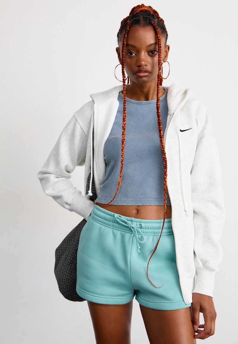 Jeune femme aux longues tresses rouges portant un sweat à capuche blanc zippé, un crop top bleu, un short turquoise et de grandes boucles d'oreilles en forme de créole, se tenant devant un fond uni.