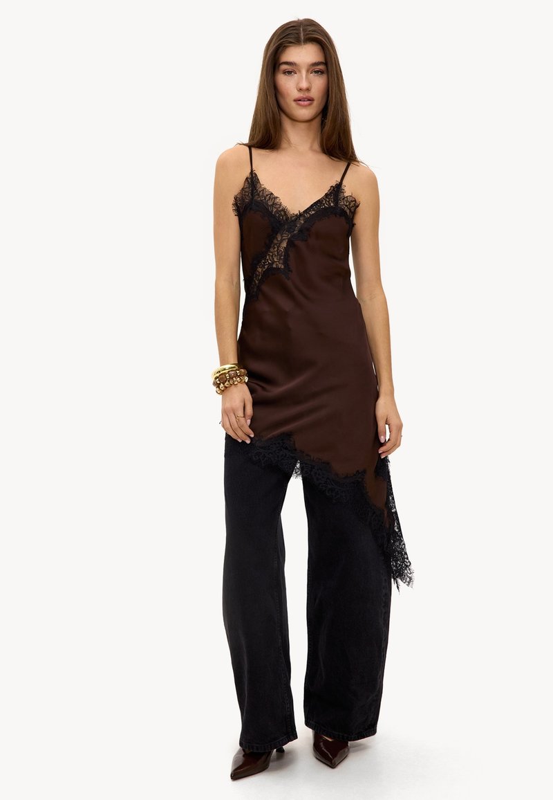 Jeune femme portant une robe nuisette asymétrique marron foncé ornée de dentelle, par-dessus un pantalon large noir, avec des chaussures pointues et des bracelets dorés.