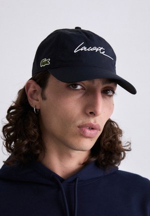 Căciulă de baseball navy cu logo-ul alb "Lacoste" scris cu litere cursive și emblema crocodilului verde. Material moale, cu bor curbat și curea ajustabilă.