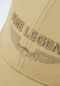 Casquette beige en tissu texturé, ornée d'un texte brodé marron "PME LEGEND" et d'accents graphiques en forme d'ailes.