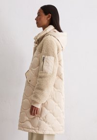 Frau trägt beige gesteppte Jacke mit Kapuze und strukturierten Fleece-Ärmeln, steht seitlich an einer schlichten weißen Wand.