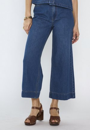 Femme portant un jean bleu large court, des sandales à talons marron, ainsi que des bracelets et des bagues en or sur un fond clair uni.