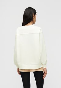 Γυναίκα με μακριά σκούρα μαλλιά, ντυμένη με άνετο λευκό φούτερ, μπεζ σορτς και μαύρες καλσόν, γυρισμένη πλάτη προς ένα απλό φωτεινό φόντο.