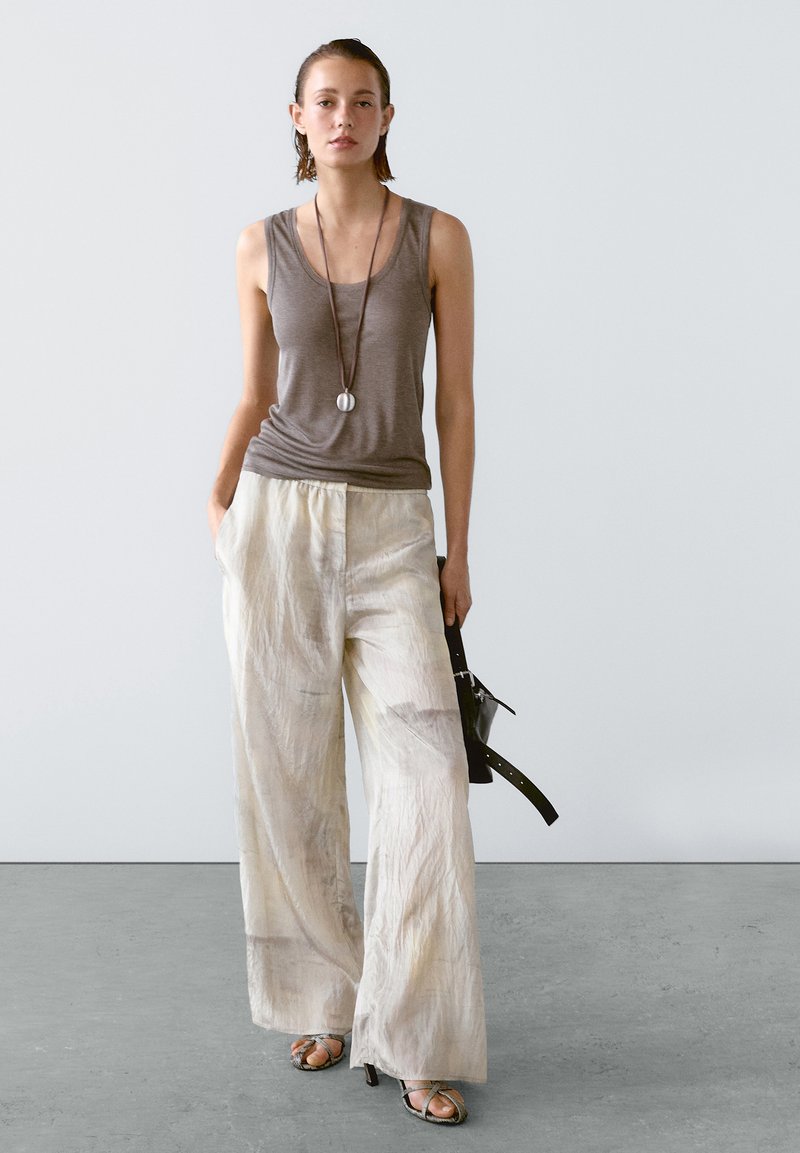 Lichtgrijze tanktop, losse crèmekleurige linnenbroek en grijze sandalen met bandjes. Lange ketting met een cirkelvormige hanger. Minimalistische stijl.