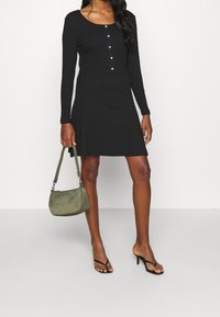 Svart långärmad klänning med rund halsringning och knäppning, tillsammans med en olivgrön handväska och svarta klacksandaler.