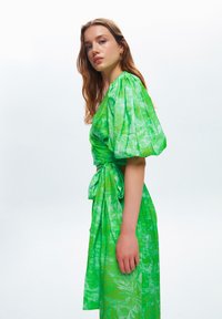 Groene jurk met poffende mouwen, een getailleerde taille en strikdetail, voorzien van een textuur met botanische print in wisselende tinten groen.