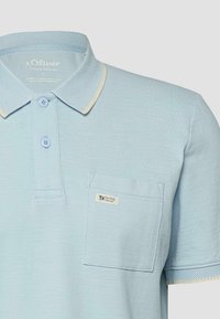Hellblaues Poloshirt mit kurzen Ärmeln, zwei Knöpfen, beigem Besatz am Kragen und einer Brusttasche mit der Aufschrift "Elevated Casual."