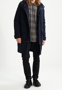 Marineblauern Wollmantel mit großer Kapuze, Frontreißverschluss und zwei Seitentaschen, getragen über einem grauen karierten Hemd, kombiniert mit schwarzen Hosen und Schuhen.