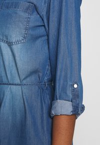 Denimmekko vaaleasta tummansiniseen liukuvärjäyksellä, jossa on rintatasku, käännetyt hihansuut ja joustava vyötäröyksityiskohta.