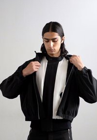 Schwarze Bomberjacke mit weißem Innenfutter, Reißverschluss vorne, elastischen Bündchen und Saum. Das Modell öffnet die Jacke und zeigt die kontrastierenden Farben.