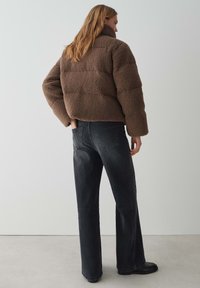 Braune Fleecejacke mit hohem Kragen, die ein puffiges Design aufweist. Kombiniert mit schwarzen weit geschnittenen Jeans und schwarzen flachen Stiefeln.