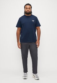 Tamsiai mėlynas medvilninis marškinėlis trumpomis rankovėmis ir mažu krūtinės logotipu, derinamas su pilkomis kargo kelnėmis su šoninėmis kišenėmis.