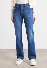 Modellen har på seg blå vide jeans og en kremfarget topp, står på en nøytral overflate. Hvite sneakers fullfører det uformelle antrekket.