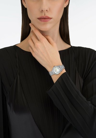 Hõbedane käekell ümmarguse näo ja kahe kuldse käega, millel on tekstuuriga hõbedane rihm. Kandjal on neutraalne ilme ja seljas on must kortsunud tops.