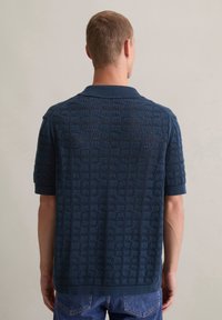 Marineblauwe gebreide polo met een textuur van geometrische patronen, korte mouwen en een contrasterende kraag. De achterkant toont een geribbelde boord en manchetten.