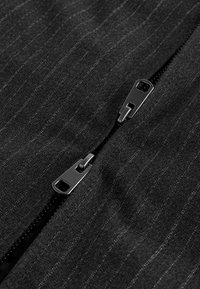 Deux curseurs de fermeture éclair en métal noir sur un tissu gris foncé à fines rayures, positionnés en diagonale et partiellement fermés.