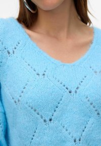 Hellblauer, gestrickter Pullover mit V-Ausschnitt, strukturierten Rautenmuster und weichem, flauschigem Material. Er ist an den Schultern verkürzt.