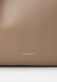 Sac en cuir texturé marron avec le logo embossé "Coccinelle" en or au centre en bas. Design simple et minimaliste.