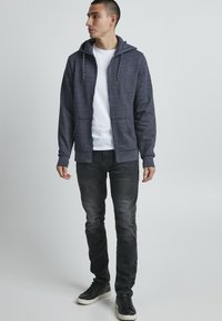 Felpa con zip di colore navy con finitura testurizzata, cappuccio con coulisse e due tasche frontali, abbinata a una T-shirt bianca e jeans scuri.