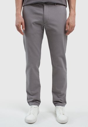 Homme portant un pantalon gris à coupe droite et des baskets blanches à lacets, debout avec les mains détendues le long du corps sur un fond uni.